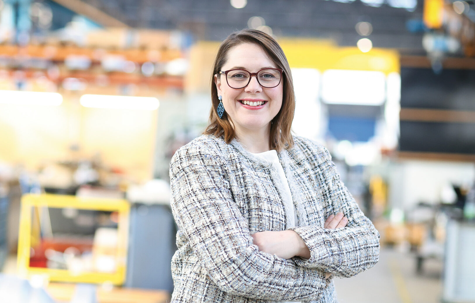 Eine Frau mit Brille und karierter Jacke lächelt mit verschränkten Armen in einem hellen, modernen Industriearbeitsraum.