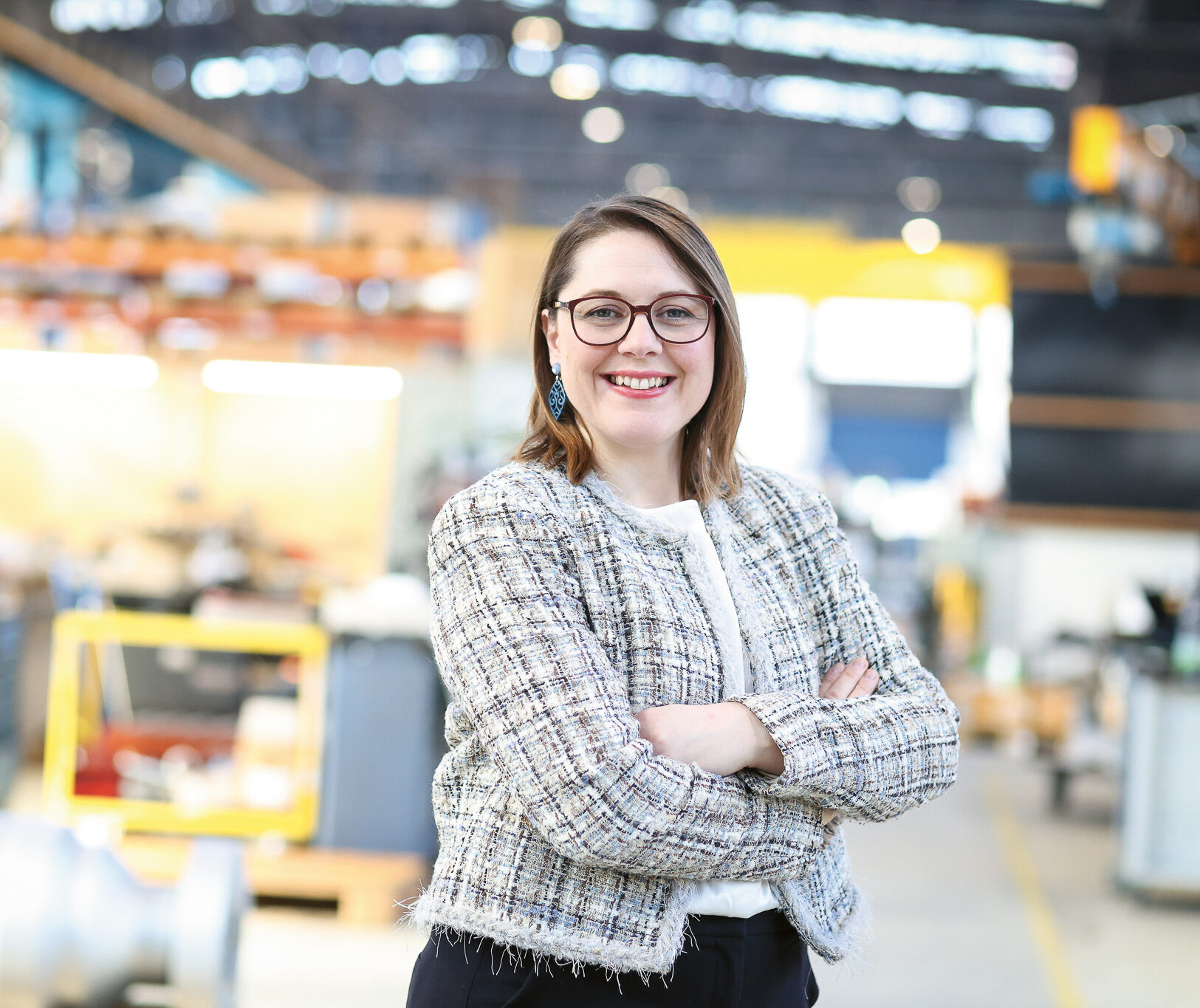 Eine Frau mit Brille und karierter Jacke lächelt mit verschränkten Armen in einem hellen, modernen Industriearbeitsraum.