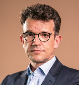 Ein Mann im Anzug und mit Brille steht vor einem schlichten Hintergrund und blickt selbstbewusst und mit neutraler Miene in die Kamera.