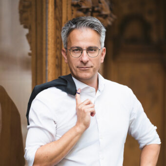 Ein Mann mit Brille und grauem Haar steht selbstbewusst mit einer Jacke über der Schulter da und spiegelt sich in einer Glaswand neben verschnörkelten Holztafeln.