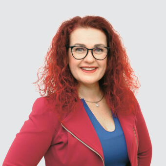 Eine lächelnde Frau mit lockigem rotem Haar und schwarzer Brille, die einen roten Blazer über einem blauen Oberteil trägt, posiert selbstbewusst vor einem hellgrauen Hintergrund.