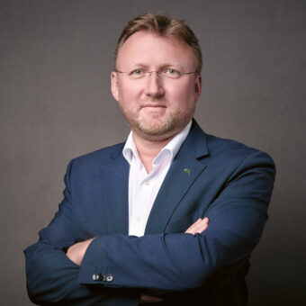 Ein Mann in blauem Anzug und weißem Hemd steht mit verschränkten Armen vor einem schlichten grauen Hintergrund vor der Kamera.