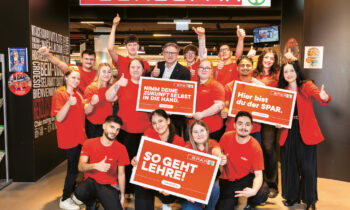 Eine Gruppe von EUROSPAR-Mitarbeitern in roten Hemden posiert lächelnd am Eingang des Ladens und wirbt mit Schildern in deutscher Sprache für Karrieremöglichkeiten.