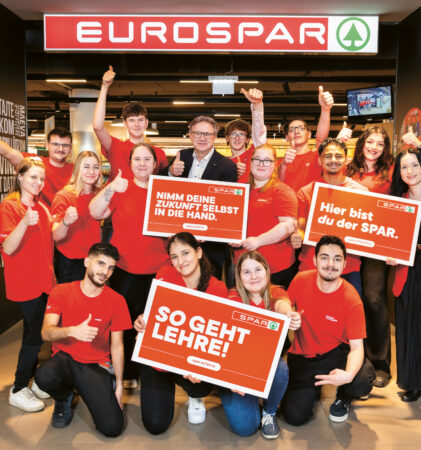 Eine Gruppe von EUROSPAR-Mitarbeitern in roten Hemden posiert lächelnd am Eingang des Ladens und wirbt mit Schildern in deutscher Sprache für Karrieremöglichkeiten.