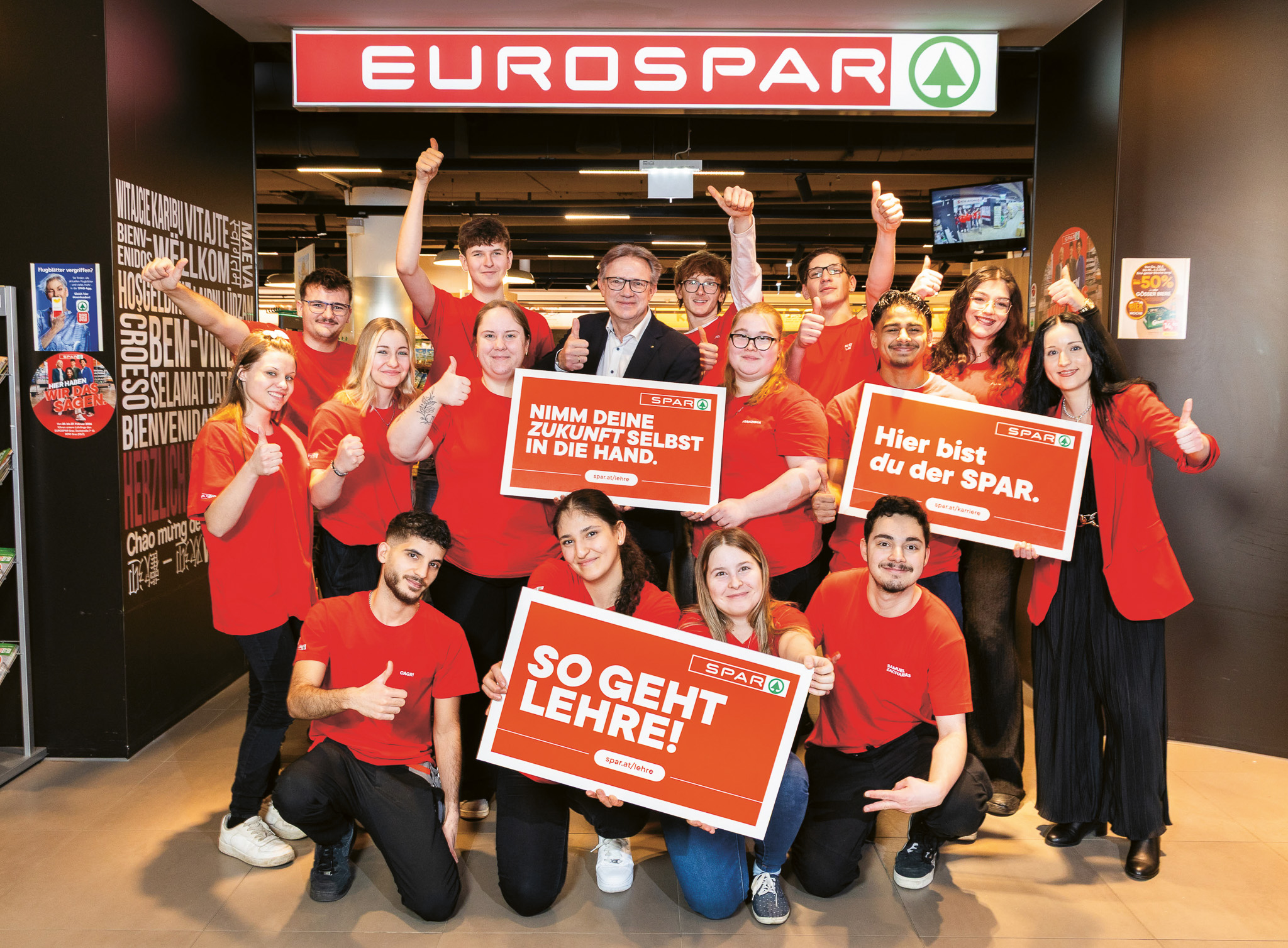 Eine Gruppe von EUROSPAR-Mitarbeitern in roten Hemden posiert lächelnd am Eingang des Ladens und wirbt mit Schildern in deutscher Sprache für Karrieremöglichkeiten.