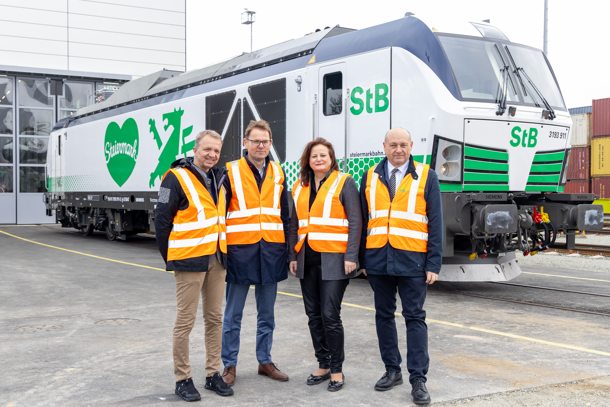 Vier Personen in orangefarbenen Sicherheitswesten stehen vor einer weiß-grünen Lokomotive mit der Aufschrift "StB" vor einem Industriegebäude.