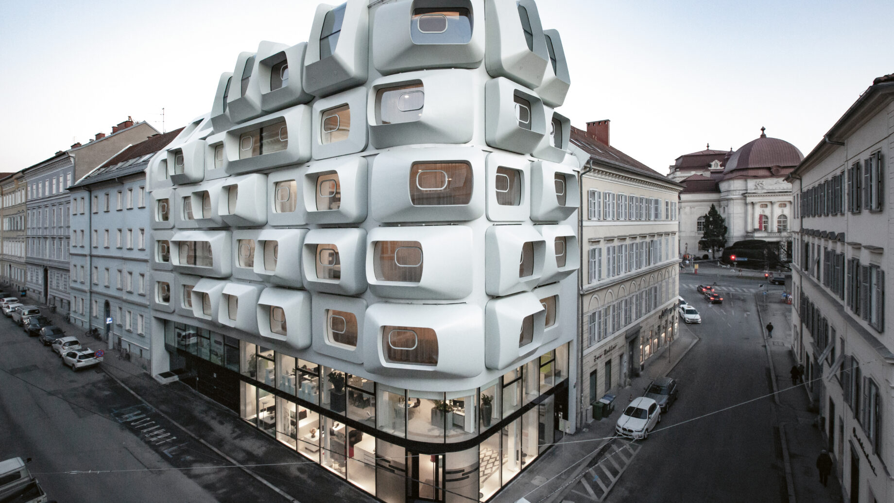 Futuristisches weißes Gebäude mit geometrischen, kastenförmigen Fenstern an einer Straßenecke, umgeben von traditioneller Architektur.