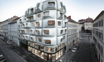 Futuristisches weißes Gebäude mit geometrischen, kastenförmigen Fenstern an einer Straßenecke, umgeben von traditioneller Architektur.