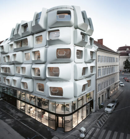 Futuristisches weißes Gebäude mit geometrischen, kastenförmigen Fenstern an einer Straßenecke, umgeben von traditioneller Architektur.