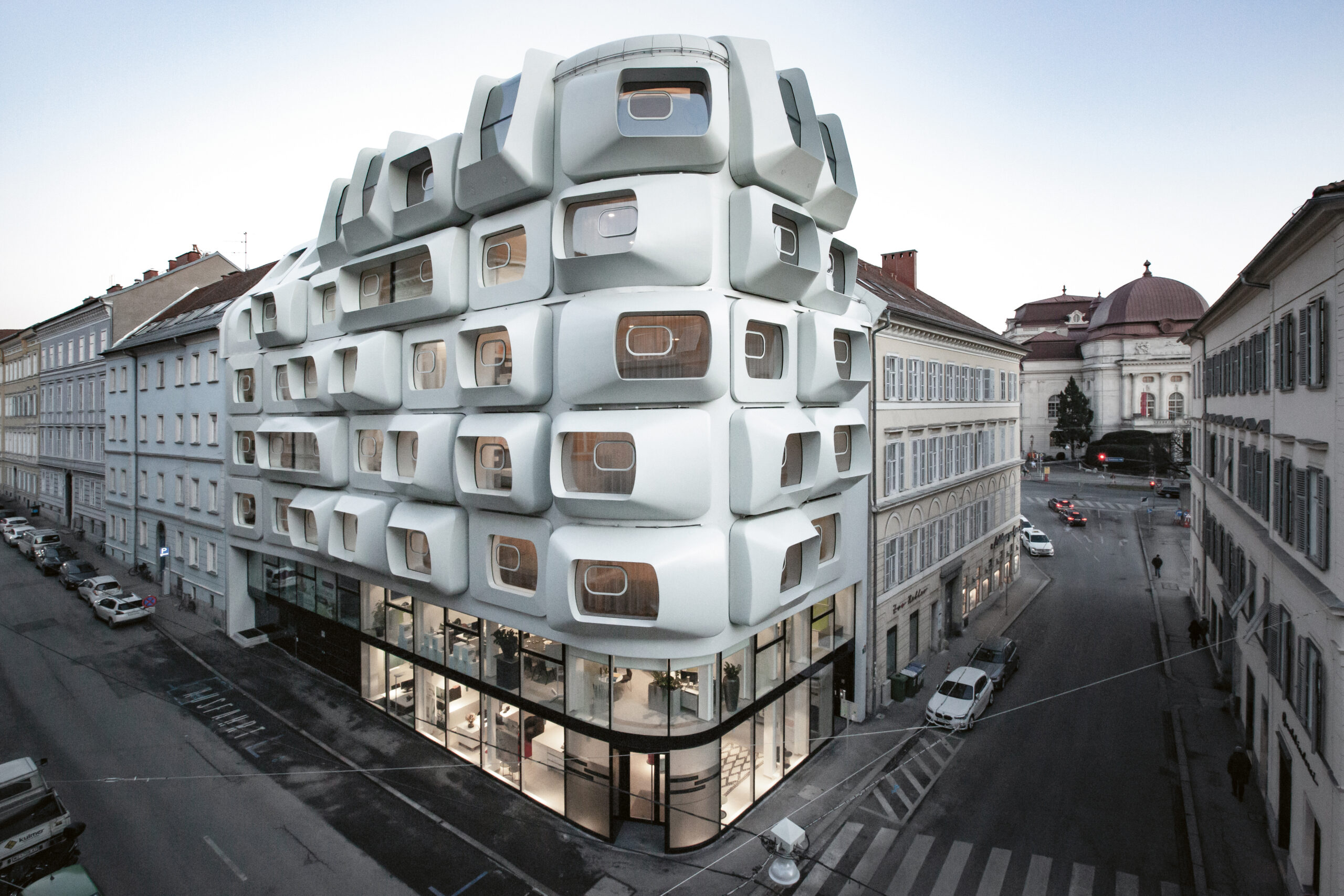Futuristisches weißes Gebäude mit geometrischen, kastenförmigen Fenstern an einer Straßenecke, umgeben von traditioneller Architektur.