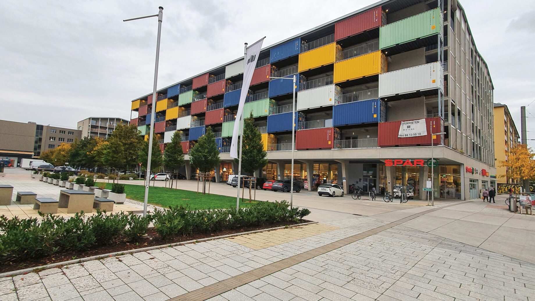 Ein modernes Gebäude aus bunt gestapelten Schiffscontainern, mit Geschäften im Erdgeschoss und einem Platz mit Bäumen und Bänken davor.
