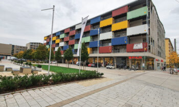 Ein modernes Gebäude aus bunt gestapelten Schiffscontainern, mit Geschäften im Erdgeschoss und einem Platz mit Bäumen und Bänken davor.