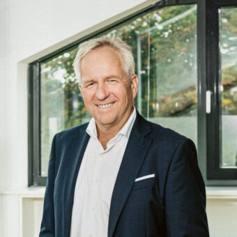 Ein lächelnder Mann in einem dunklen Anzug steht in einem modernen Büro mit großen Fenstern, die Hände in den Taschen.