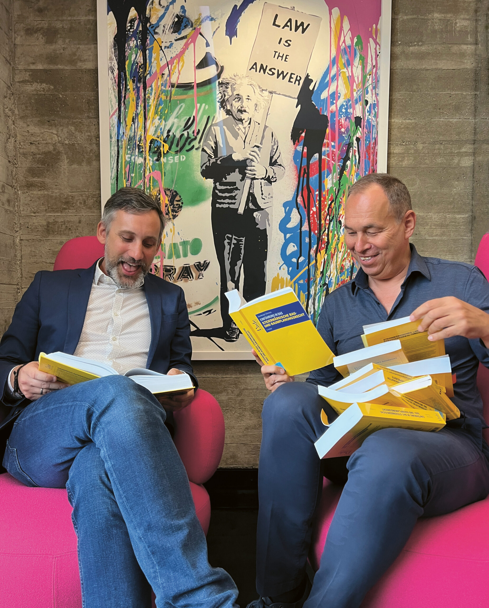 Zwei Männer sitzen auf rosa Stühlen, lächeln und lesen gelbe Bücher. Hinter ihnen ist eine bunte Kunstfigur zu sehen, die ein Schild mit der Aufschrift "GESETZ IST DIE ANTWORT" hält.