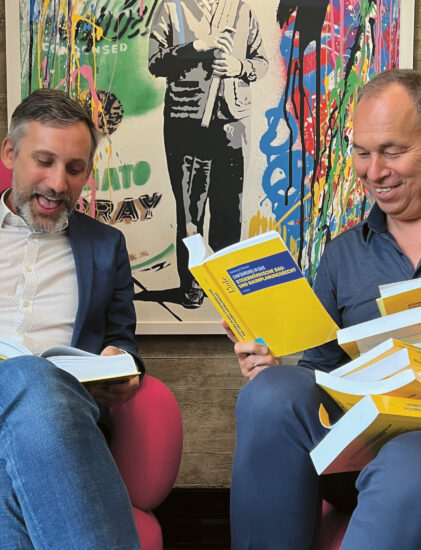 Zwei Männer sitzen auf rosafarbenen Stühlen und lesen lächelnd in gelben Gesetzesbüchern, während hinter ihnen ein buntes Kunstwerk mit dem Titel "Law is the Answer" an der Wand hängt.