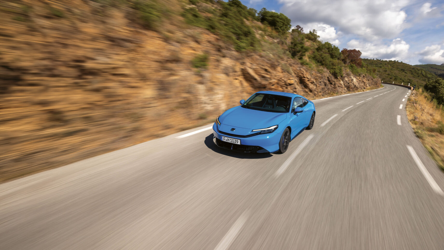 Ein blaues Auto fährt zügig über eine kurvenreiche Bergstraße, die von felsigen Hügeln und Grünflächen gesäumt ist, unter einem teilweise bewölkten Himmel.