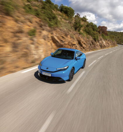 Ein blaues Auto fährt zügig über eine kurvenreiche Bergstraße, die von felsigen Hügeln und Grünflächen gesäumt ist, unter einem teilweise bewölkten Himmel.