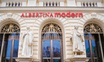 Fassade des modernen Museums Albertina mit zwei weißen Statuen und verzierten Bogenfenstern, rotes Schild über dem Eingang.