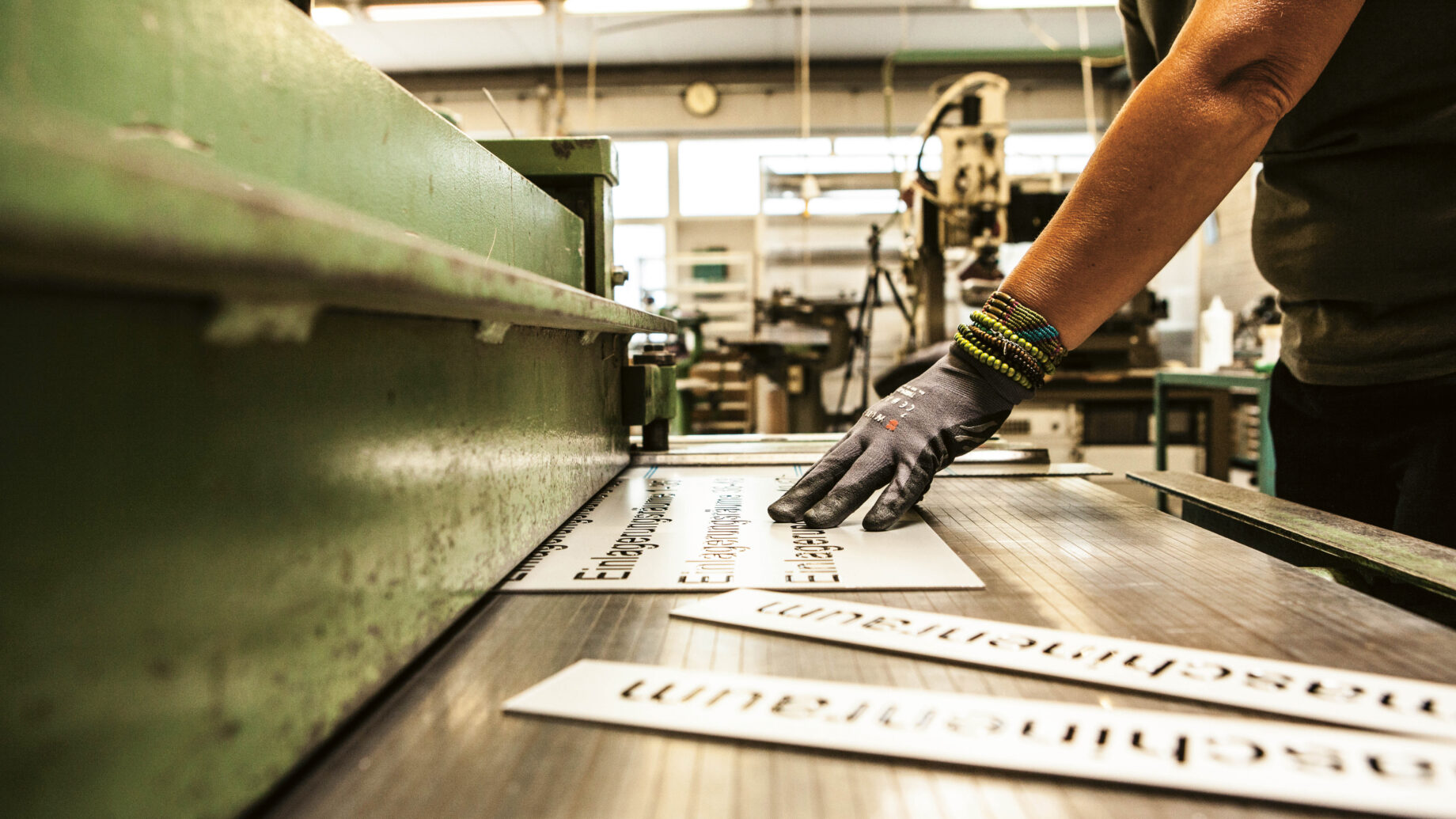 Eine Person mit Handschuhen ordnet bedruckte Schilder auf einer Schneidemaschine in einer Werkstatt mit Industrieanlagen im Hintergrund an.