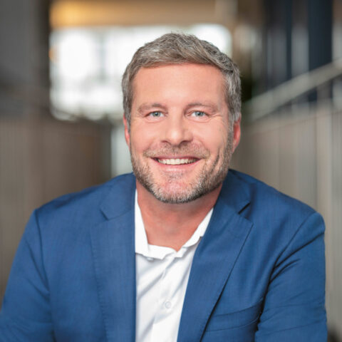 Lächelnder Mann mit kurzen grauen Haaren und Bart, der eine blaue Anzugsjacke und ein weißes Hemd trägt, sitzt in einem Haus mit unscharfem Hintergrund.
