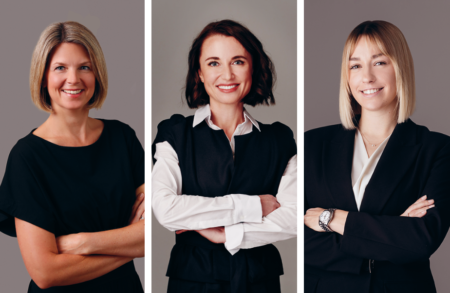 Drei Frauen in Business-Kleidung stehen mit verschränkten Armen vor einem neutralen Hintergrund und lächeln in die Kamera.