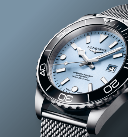 Eine silberne Longines HydroConquest Uhr mit hellblauem Zifferblatt, schwarzer Lünette, Datumsanzeige und Mesh-Metallarmband auf blauem Hintergrund mit Farbverlauf.