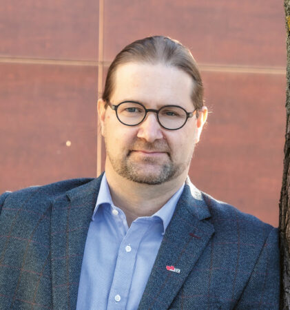 Ein Mann mit Brille und Anzugjacke lehnt an einem Baum, steht vor einer rötlich-braunen Wand und schaut in die Kamera.
