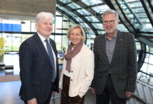 Drei ältere Erwachsene in Business-Kleidung lächeln in einem Innenraum mit moderner Glasarchitektur und natürlichem Licht - in der Nähe der Kältekammer Graz Altstadt.