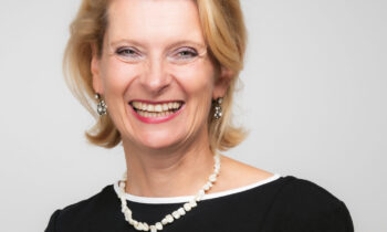 Lächelnde Frau mit blondem Haar, die eine Brille auf dem Kopf trägt, ein schwarzes Kleid, eine Perlenkette und eine schwarze Uhr, die vor einem schlichten Hintergrund steht.