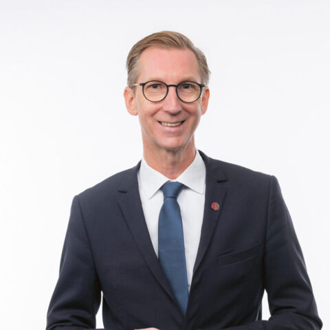 Ein Mann in einem dunklen Anzug, mit blauer Krawatte und Brille lächelt, während er vor einem schlichten weißen Hintergrund steht.