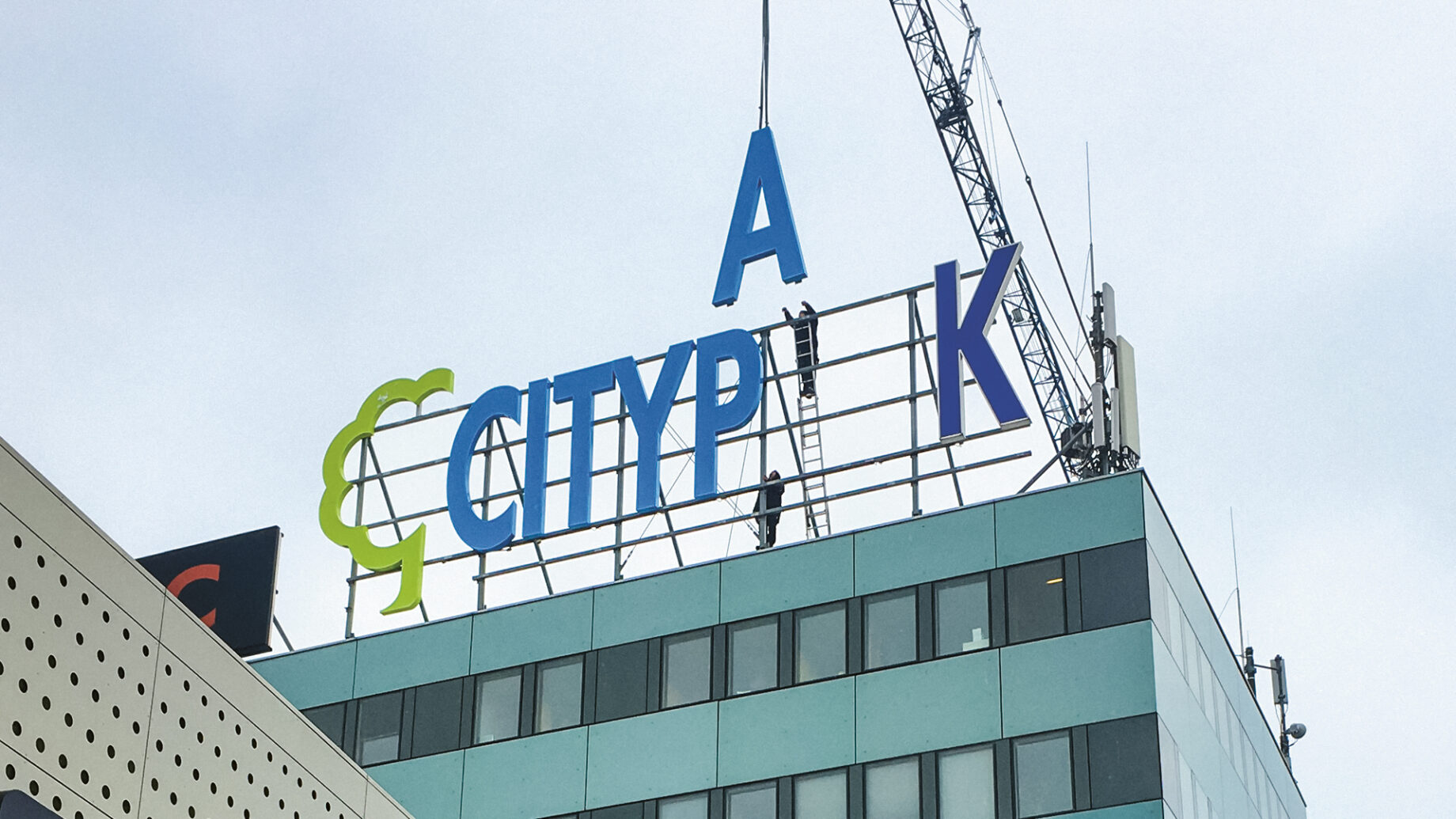 Große blaue Buchstaben mit dem Schriftzug "CITYPAK" und ein grünes Logo werden mit Hilfe eines Krans auf einem modernen Gebäude angebracht, über dem ein bewölkter Himmel hängt.