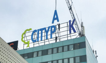 Große blaue Buchstaben mit dem Schriftzug "CITYPAK" und ein grünes Logo werden mit Hilfe eines Krans auf einem modernen Gebäude angebracht, über dem ein bewölkter Himmel hängt.