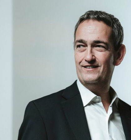 Ein Mann in einem dunklen Anzug und weißem Hemd steht in einem Haus, lächelt und blickt nach links vor einem schlichten hellen Hintergrund.