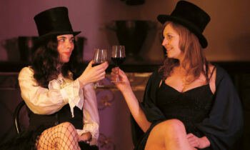 Zwei Frauen mit Zylinder und Vintage-Outfit lächeln und stoßen mit Weingläsern an, während sie in einem Haus sitzen.