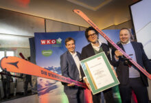 Drei Männer in Anzügen lächeln, halten lange orangefarbene Skier und eine Steiermarkbahn-Vectron-Dual-Mode-Urkunde bei einer Indoor-Skiverleihung in der Hand.