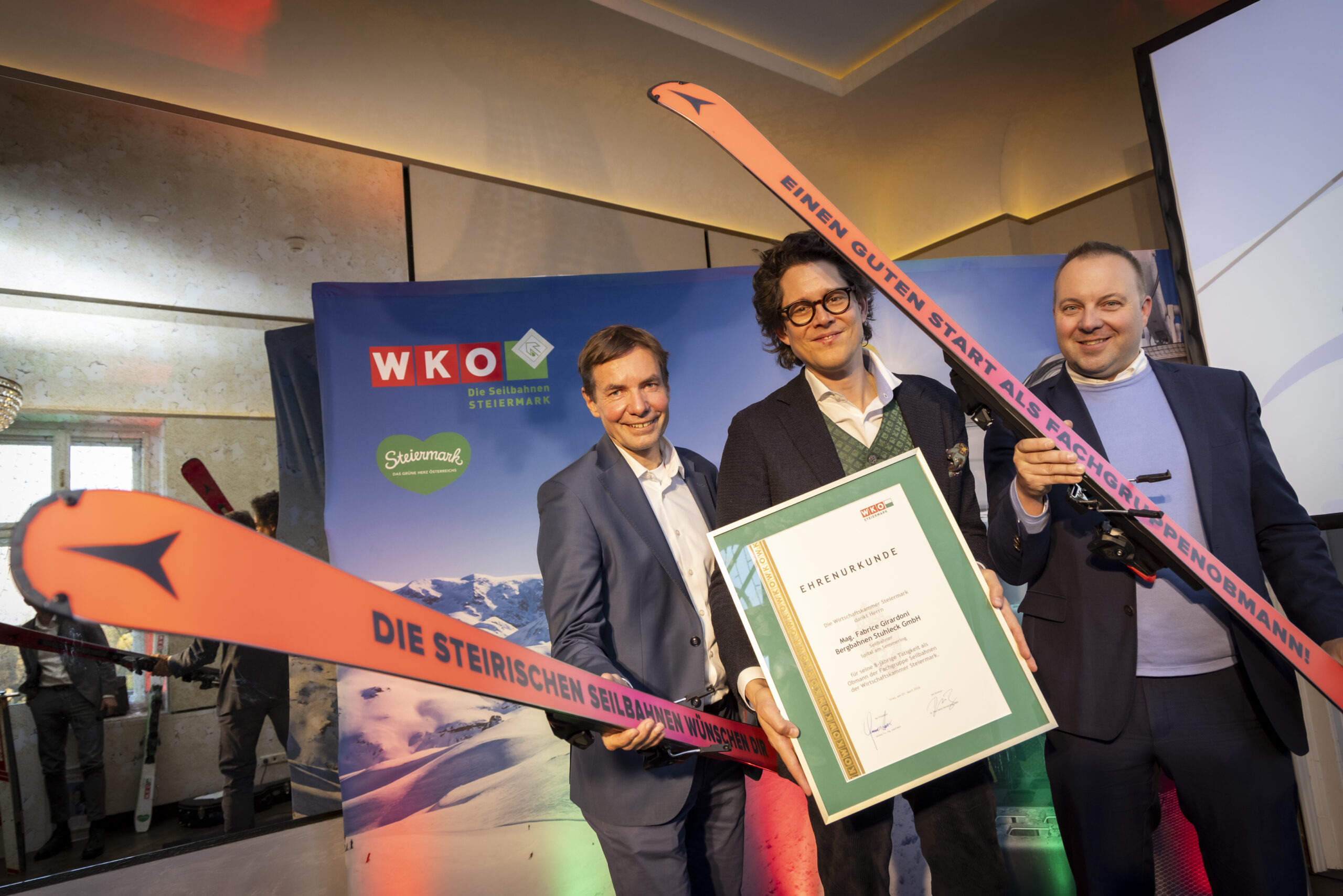 Drei Männer in Anzügen lächeln, halten lange orangefarbene Skier und eine Steiermarkbahn-Vectron-Dual-Mode-Urkunde bei einer Indoor-Skiverleihung in der Hand.