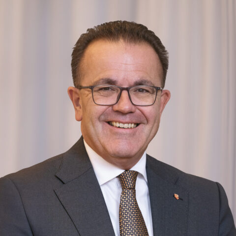 Lächelnder Mann in Anzug und Krawatte mit Brille, der vor weißen Vorhängen steht.