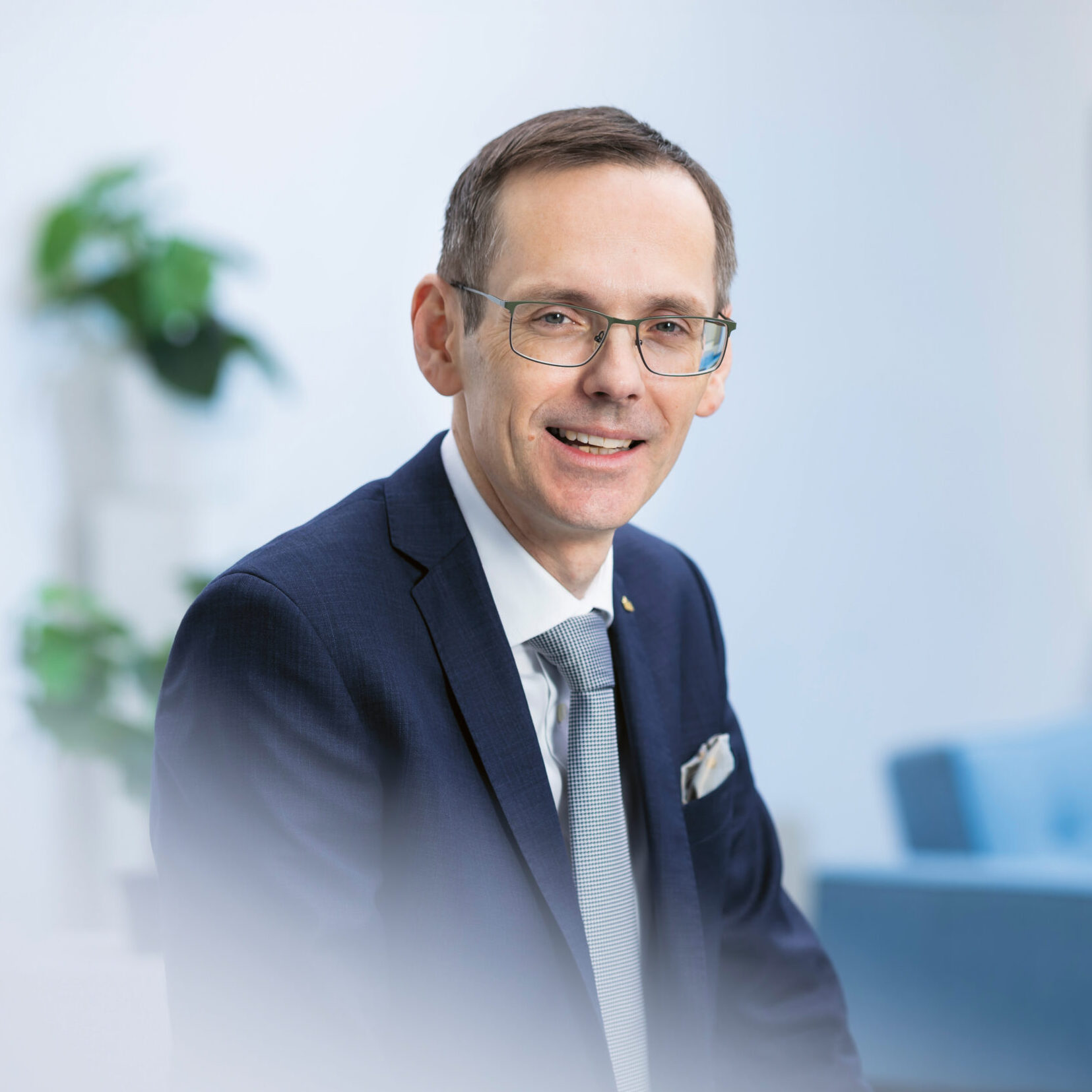 Ein Mann in Anzug und Krawatte, der eine Brille trägt, lächelt in einem hellen Büro mit blauen Möbeln und Pflanzen im Hintergrund.