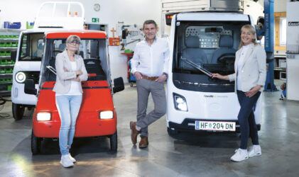 Drei Personen stehen in einer hellen Garage zwischen einem kleinen roten Fahrzeug und einem kleinen weißen Fahrzeug und lächeln in die Kamera.