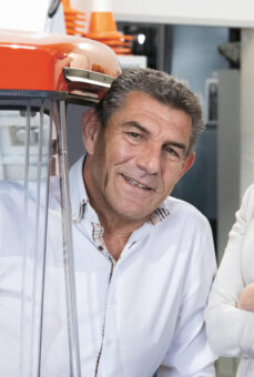 Drei lächelnde Erwachsene, zwei Frauen und ein Mann, posieren zusammen in einer Garage zwischen zwei Fahrzeugen und tragen helle Business Casual Kleidung.