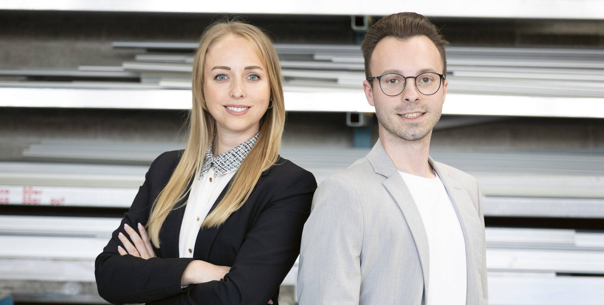 Zwei junge Berufstätige in Geschäftskleidung stehen selbstbewusst in einer industriellen Umgebung mit Metallregalen und Blechen im Hintergrund.