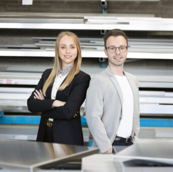 Zwei junge Berufstätige in Geschäftskleidung stehen selbstbewusst in einer industriellen Umgebung mit Metallregalen und Blechen im Hintergrund.