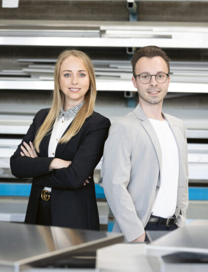 Zwei junge Berufstätige in Geschäftskleidung stehen selbstbewusst in einer industriellen Umgebung mit Metallregalen und Blechen im Hintergrund.