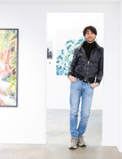 Ein Mann in schwarzer Jacke und Jeans steht in einer hellen Kunstgalerie, umgeben von bunten Gemälden an weißen Wänden.