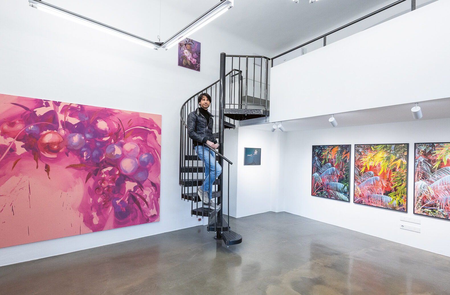 Eine Person steht auf einer Wendeltreppe in einer modernen Kunstgalerie mit farbenfrohen abstrakten und naturbezogenen Gemälden an weißen Wänden.