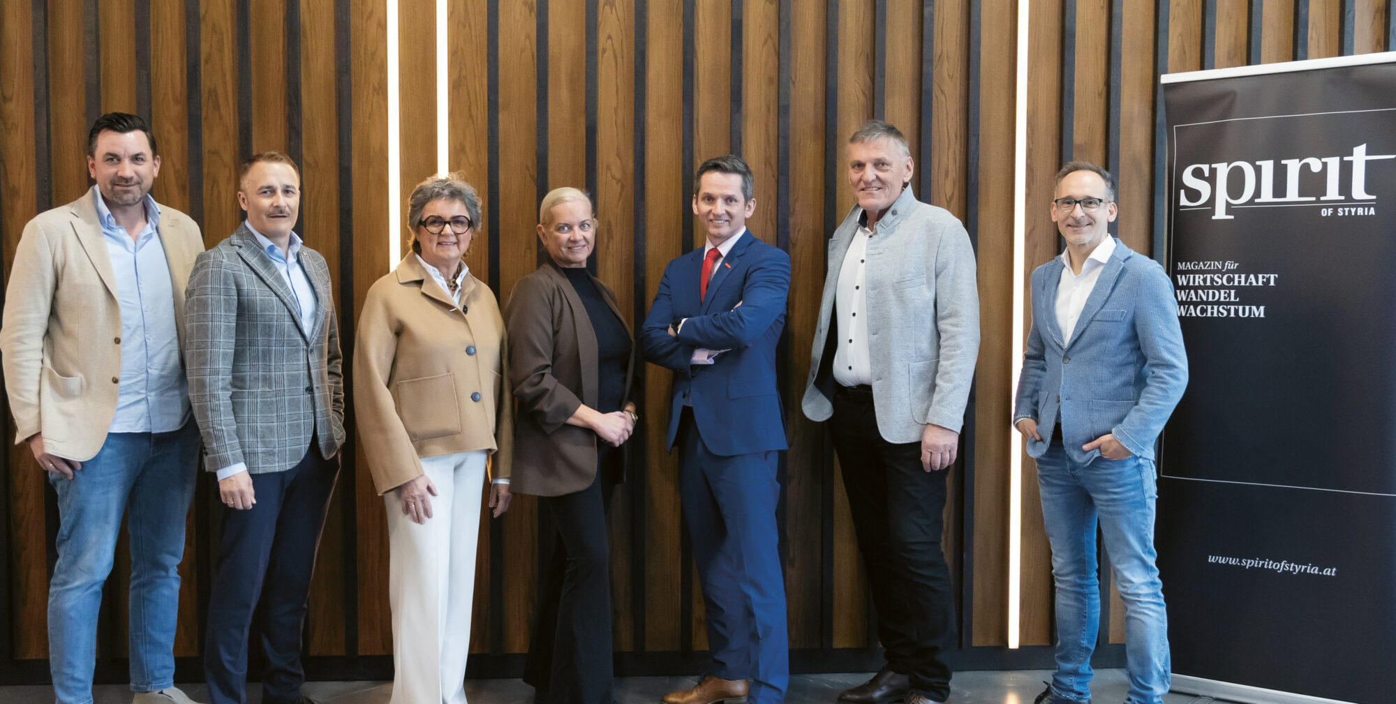 Sieben professionell gekleidete Menschen stehen lächelnd in einer Reihe neben einem "Spirit of Styria"-Banner und einer Holztafelwand.