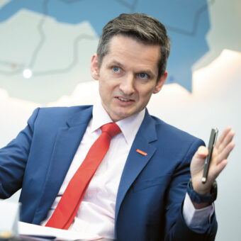 Ein Mann in blauem Anzug und roter Krawatte gestikuliert mit den Händen, während er spricht. Er sitzt an einem Tisch mit einer Landkarte an der Wand hinter ihm.