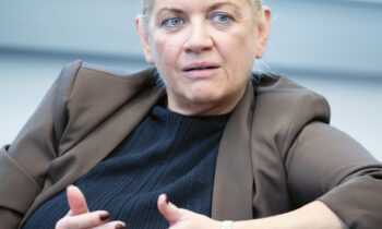 Eine Frau mit hochgestecktem blondem Haar, die einen braunen Blazer und ein schwarzes Oberteil trägt, gestikuliert mit den Händen, während sie in einer Büroumgebung spricht.