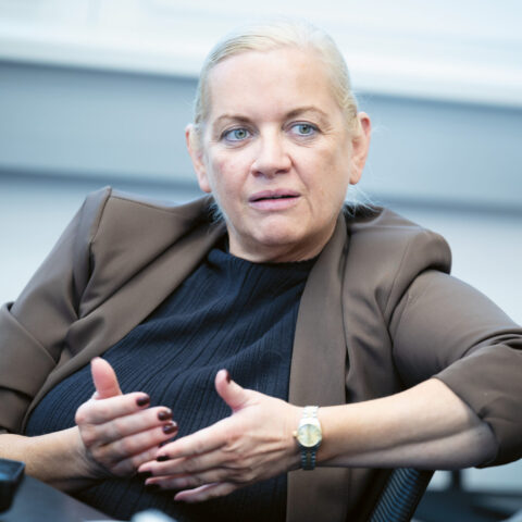 Eine Frau mit hochgestecktem blondem Haar, die einen braunen Blazer und ein schwarzes Oberteil trägt, gestikuliert mit den Händen, während sie in einer Büroumgebung spricht.