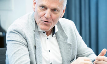 Ein älterer Mann in einem hellgrauen Blazer spricht, während er an einem Tisch sitzt und mit den Händen während einer Besprechung in einem modernen Büro gestikuliert.